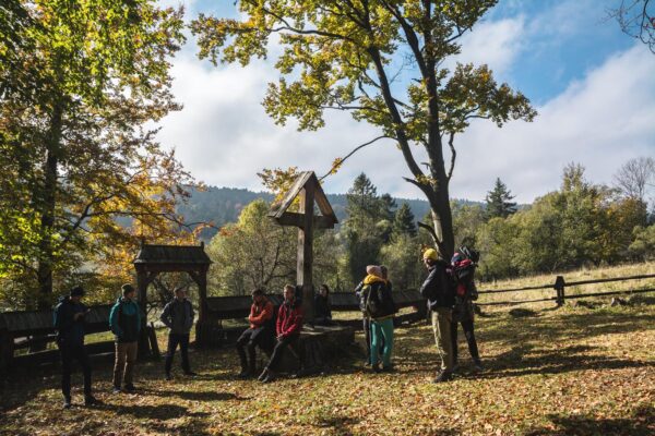 Grupa ludzi w jesiennym krajobrazie stoi przy drewnianym ogrodzeniu cerkwii.